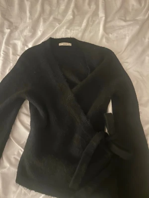 Svart omlottkofta från Gina Tricot - Mysig svart omlottkofta från Gina Tricot med knytband i midjan. Koftan har lång ärm och är tillverkad i ett mjukt, fluffigt material som ger en avslappnad men ändå stilren look. Perfekt att slänga över en topp för extra värme.