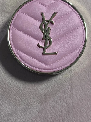 YSL rosa puderrouge i lyxig dosa - Lyxigt puderrouge från Yves Saint Laurent i en rund dosa med quiltad, ljusrosa läderlook och ikoniska YSL-loggan i metall. Rouget har en mjuk, kall rosa ton och pressat YSL-mönster. Perfekt för dig som vill ha en elegant touch i sminkväskan.