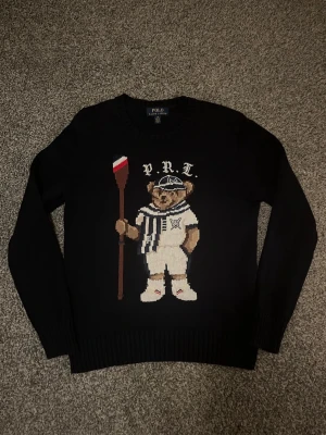 Mörkblå stickad tröja Polo Ralph Lauren - Mörkblå stickad tröja från Polo Ralph Lauren med ikonisk björn på bröstet, klädd i vit outfit och keps samt håller en åra. Rund halsringning och ribbade muddar. Perfekt för dig som vill ha en klassisk men lekfull look. Storlek L ungdom passar liten xs