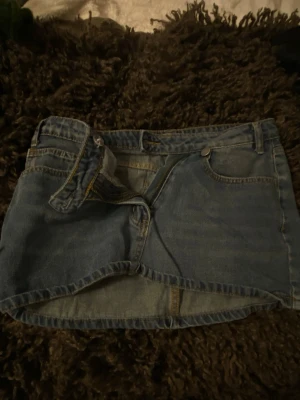 Blå jeanskjol med dragkedja - Säljer en klassisk blå jeanskjol i kort modell med dragkedja och knapp framtill. Kjolen har fickor både fram och bak samt bälteshällor. Perfekt för dig som gillar en enkel och tidlös denimlook.