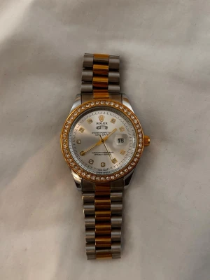 Klocka  - Säljer en stilren Rolle Datejust med tvåtonad länk i rostfritt stål och guld. Urtavlan är silverfärgad med datumvisning och gulddetaljer, samt en bezel dekorerad med gnistrande stenar. Klassisk och lyxig design som sticker ut på handleden. Batteri driven som just nu är död men kan enkelt bytas ut.