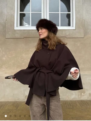Mörkbrun cape-kappa med knytband - Stilren mörkbrun cape-kappa med hög krage, knappar framtill och brett knytband i midjan. Kappan har vida ärmar och en elegant, lös passform. Perfekt för lager-på-lager och ger en trendig siluett. Materialet ser ut att vara ullblandning.