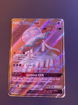 Nihilego GX Pokémonkort Ultra Beast - Säljer ett holografiskt Nihilego GX Ultra Beast Pokémonkort med 180 HP. Kortet har färger i lila, blått och silver med skimrande effekt. Det är en samlarkort från Sun & Moon-serien och har attacker som Lock Up och Symbiont GX.