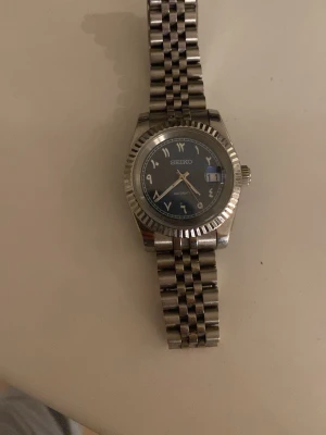 Seiko klocka - Snygg Seiko automatisk klocka med blå urtavla och datumfönster. Klockan har ett klassiskt rundat boett i rostfritt stål och matchande länkarmband. Indexen är i silver och urtavlan har en stilren look med tydliga siffror och visare.