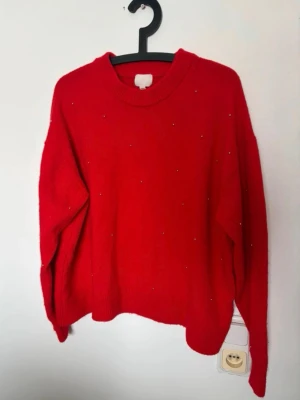 Röd stickad tröja från H&M - Snygg röd stickad tröja från H&M med rund halsringning och små diskreta pärldetaljer över hela tröjan. Tröjan har en avslappnad passform och ribbade muddar vid hals, ärmslut och nederkant. Perfekt för att addera färg till din outfit.