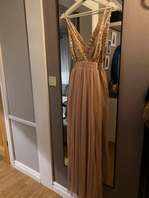 Champagnefärgad långklänning med paljetter - Säljer en elegant långklänning i champagnefärg med glittrig paljettöverdel och djup v-ringning. Kjolen är i mjukt, tunt tyg som faller snyggt och ger en romantisk look. Klänningen har smala axelband och markerad midja.