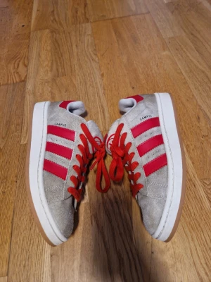 Adidas Campus sneakers beige och röd - Säljer ett par Adidas Campus sneakers i beige mocka med röda detaljer och snörning. Skorna har de klassiska tre ränderna i rött på sidorna och en vit sula med gummisula undertill. Perfekt för dig som gillar retrostil och vill sticka ut med färg.