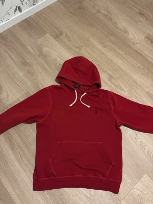 Röd hoodie från Polo Ralph Lauren - Snygg röd hoodie från Polo Ralph Lauren med klassisk huva och vita dragsnören. Har en känguruficka framtill och den ikoniska lilla loggan broderad på bröstet. Tillverkad i mjukt bomullsmaterial som känns skönt mot huden. Perfekt för en avslappnad och stilren look.