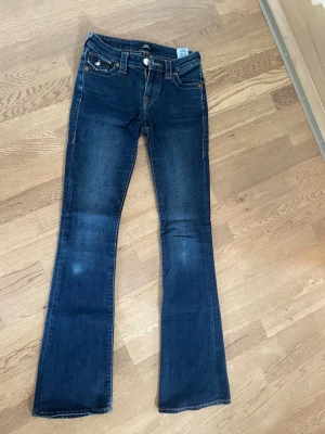 Mörkblå bootcut jeans från True Religion - Snygga mörkblå jeans från True Religion med bootcut passform och klassiska kontrastsömmar. Jeansen har dekorativa bakfickor med lock och knappar samt detaljerade sömmar. Tillverkade i jeansmaterial med en skön stretch. Jeansen är lite slitna vid knäna (se bild) men annars är de i toppenskick!