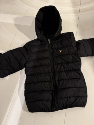 Svart dunjacka från Lyle & Scott - Svart puffer dunjacka från Lyle & Scott med huva och dragkedja framtill. Jackan har det klassiska guldiga logomärket på bröstet och quiltad design. Perfekt för kalla dagar och riktigt snygg till vardagslooken.har ett litet jack under märket går att sy.