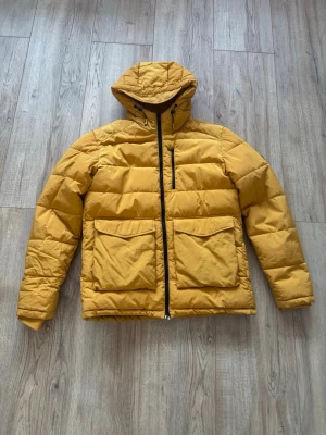Jack & Jones Vinterjacka (Stl L) – Vattenavvisande & Varm - Säljer en gul jacka från Jack & Jones (Originals) i storlek Large. En rejäl och funktionell vinterjacka som håller både värme och stil.  Egenskaper & Detaljer: • Märke: Jack & Jones Originals. • Storlek: Large (L). • Färg: Gul • Water Repellent: Materialet är vattenavvisande. • Storm Cuff: Inre muddar i ärmsluten som stänger ute vind och kyla. • Adjustable Hood: Justerbar huva för bra passform. • Handwarmer Pockets: Praktiska fickor framtill för att värma händerna.  Perfekt jacka för hösten!