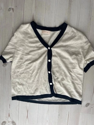 Kortärmad kofta från Torio - Stilren kortärmad kofta från Torio i ljusbeige med mörkblå kantband och knappar framtill. Koftan har v-ringning och en avslappnad passform, perfekt för lager-på-lager. Materialet känns mjukt och lätt.