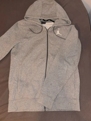 Grå Jordan hoodie med dragkedja - Snygg grå hoodie från Jordan med hel dragkedja och klassisk Jumpman-logga broderad på bröstet. Mjuk insida och justerbar huva med dragsko. Perfekt för en sportig och avslappnad stil.
