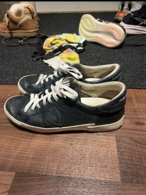 Mörkblå sneakers från Philippe Model - Stilrena svarta sneakers från Philippe Model med vita snören och vit sula. Skorna har en klassisk låg profil och är tillverkade i skinn med diskreta detaljer på sidan. Perfekta för dig som gillar en clean och tidlös look.