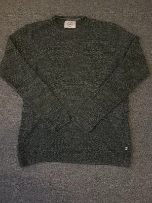 Mörkgrå stickad tröja Kronstadt - Snygg mörkgrå stickad tröja från Kronstadt med rund halsringning och diskret melerad struktur. Tröjan har långa ärmar och ribbade muddar vid ärmslut och nederkant. Perfekt för lager på lager och enkel att matcha med jeans.