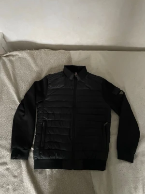 Svart cardigan från Guess - Stilren svart jacka från Guess med quiltade partier och dragkedja framtill. Jackan har långärmade ärmar, ribbade muddar och klassisk krage. Tillverkad i polyester och syntetmaterial för en lätt och bekväm känsla.