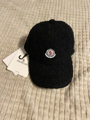 Svart teddykeps från Moncler - Snygg svart keps från Moncler i teddy-material med klassisk böjd skärm och broderad logotyp framtill. Perfekt accessoar för dig som vill sticka ut med en trendig och exklusiv look. Kepsen har justerbar passform och mjuk textur.