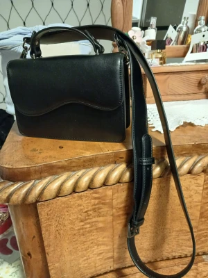 Svart axelväska i skinn - Snygg svart axelväska i fejkskinn med stilren design och justerbar axelrem. Väskan har ett lock med magnetknäppning och ett praktiskt innerfack. Perfekt storlek för det viktigaste och passar till många olika outfits.