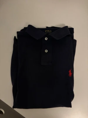 Mörkblå pikétröja från Ralph Lauren - Säljer en mörkblå pikétröja från Ralph Lauren med klassisk krage och knappar framtill. Tröjan har den ikoniska röda logotypen broderad på bröstet och är tillverkad i mjuk bomull. Perfekt för en clean och stilren look.Den är lite solblekt.Men inget man tänker på.