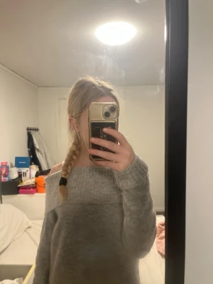 Grå off shoulder stickad tröja - Mysig grå stickad tröja med off shoulder-design och bred ribbad kant upptill. Tröjan har långa ärmar och är gjord i ett mjukt, fluffigt material som passar perfekt för kyliga dagar. Enkel att styla med jeans eller kjol för en avslappnad look.