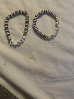 Två färgglada bokstavsarmband - Säljer två elastiska armband med bokstavspärlor. Ena armbandet har pärlor i vitt, turkost, rosa och guld, och bokstäverna KNA. Det andra har lila, vita och guldfärgade pärlor med bokstäverna SHIN. Perfekta för dig som gillar personliga accessoarer.