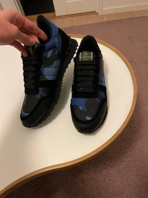 Valentino Rockrunner blå sneakers - Snygga Valentino Rockrunner sneakers i blått och svart med coolt kamouflagemönster. Skorna har svarta snören, platt sula och detaljer i skinn och textil. Perfekta för dig som vill sticka ut med stilrena och trendiga sneakers. 