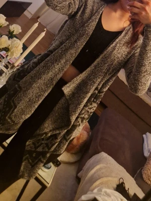 Grå lång kofta med mönster - Lång och mysig grå kofta med svart och vitt zigzag-mönster nedtill. Koftan har öppen front utan knappar och långa ärmar. Perfekt att slänga över en topp för en avslappnad look.