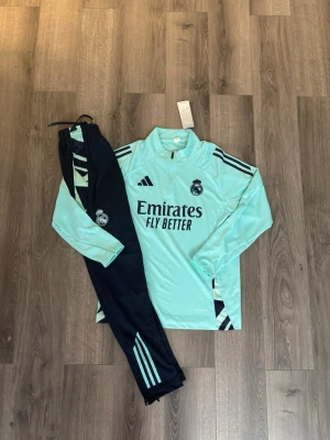 Real Madrid träningsset Adidas mint - Säljer ett Real Madrid träningsset från Adidas i mintgrönt och svart. Tröjan har halv dragkedja, svarta ränder på axlarna och klubbmärke på bröstet. Byxorna är svarta med mintgröna detaljer och klubbmärke. Materialet är funktionspolyester, perfekt för träning. Storleken är ca 164 cm+