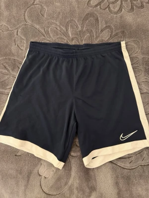 Svarta Nike shorts med vita detaljer - Snygga svarta shorts från Nike med vita kanter längs sidorna och nederkanten. Klassisk Nike-logga framtill. Perfekta för träning eller sportiga dagar. Tillverkade i ett lätt och snabbtorkande syntetmaterial med elastisk midja för skön passform.