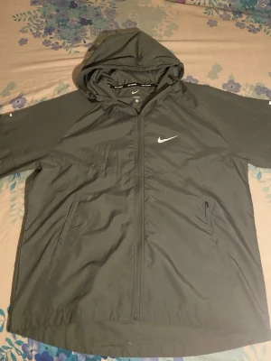 Nike Miller Grey vindjacka  - Snygg grå vindjacka från Nike med dragkedja framtill och två sidofickor. Jackan har en praktisk huva och den klassiska vita Nike-loggan på bröstet. Tillverkad i lätt och vindtätt material, perfekt för blåsiga dagar. Helt oanvänd!