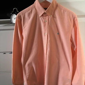 Aprikosfärgad skjorta från Morris - Snygg aprikosfärgad skjorta från Morris med klassisk button down-krage och vita knappar. Skjortan har en broderad blå logga på bröstet och är tillverkad i mjuk bomull. Perfekt för dig som gillar stilrena och fräscha plagg.