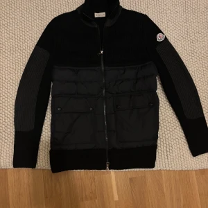 Svart stickad pufferjacka Moncler - Svart pufferjacka från Moncler med stickade partier och quiltad front. Jackan har ribbade muddar, två fickor med knappar och dragkedja hela vägen. Moncler-logga på ärmen och cool kontrast mellan material och struktur. Perfekt för dig som gillar street och premium.