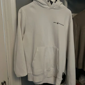 Vit hoodie från Sail Racing - Vit hoodie från Sail Racing med svart logga på bröstet. Klassisk modell med huva och snörning samt stor magficka framtill. Perfekt för dig som gillar en clean och sportig look.