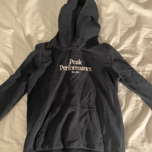 Mörkblå hoodie från Peak Performance - Säljer en mörkblå hoodie från Peak Performance med vit broderad logga på bröstet. Tröjan har huva, magficka och långa ärmar. Perfekt för dig som gillar en enkel och stilren look.