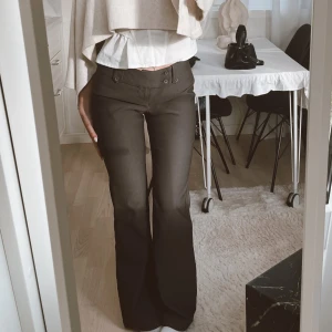 Svarta bootcut byxor med fickor - Snygga svarta byxor med bootcut-ben och låg midja. Byxorna har dubbla knappar framtill och fickor bak med lock. Perfekta för dig som gillar en klassisk men trendig look. 