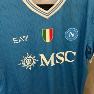 Napoli EA7 blå fotbollströja - Napoli hemmatröja från EA7 i blått med vita detaljer och klubbmärke på bröstet. Tröjan har V-ringning, korta ärmar och tryckta sponsorer. Materialet är lätt och andas, perfekt för match eller träning. Diskret mönster med N-loggor över hela tyget.