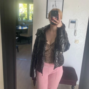 Svart pufferjacka med dragkedja - Säljer en svart, glansig pufferjacka med quiltad design och hög krage. Jackan har två fickor med dragkedja och en snygg, lite boxig passform. Perfekt för dig som vill ha en varm och stilren jacka med streetkänsla.