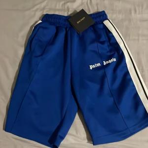 Blå Palm Angels shorts med vit rand - Säljer ett par blå shorts från Palm Angels med vit rand längs sidorna och logga broderad på vänster ben. De har elastisk midja och är tillverkade i 100% polyester, perfekta för en sportig och trendig look.
