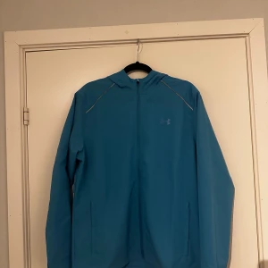 Blå vindjacka från Under Armour - Snygg blå vindjacka från Under Armour med huva och hel dragkedja. Jackan har diskreta reflexdetaljer på axlarna, två fickor framtill och stor logga på ryggen. Tillverkad i lätt och ventilerande syntetmaterial, perfekt för träning utomhus.