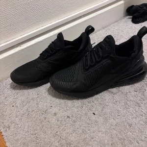 Svarta Nike Air Max 270 sneakers - Säljer ett par helsvarta Nike Air Max 270 sneakers med snygg, sportig design och synlig luftbubbla i hälen. Skorna har mesh-överdel för bra andningsförmåga och klassisk snörning. Perfekta för dig som gillar stilrena och bekväma sneakers.