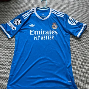 Real Madrid Mbappé blå matchtröja - Blå Real Madrid fotbollströja från Adidas med Mbappé och nummer 9 på ryggen. Tröjan har vita detaljer, klubbmärke, Champions League-märke och sponsortryck. Tillverkad i lätt och ventilerande material, perfekt för match eller träning.