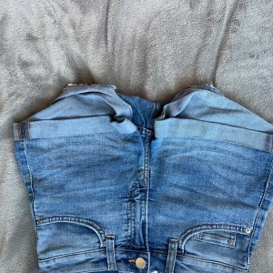Blå jeansshorts från H&M - Säljer ett par klassiska blå jeansshorts från H&M i storlek 32. De har hög midja, uppvikta benslut och femficksdesign. Perfekta för en chill sommarlook och lätta att matcha med allt. Snygga detaljer med synliga sömmar och bälteshällor.