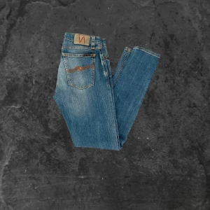 Blå jeans från Nudie Jeans - Säljer ett par klassiska blå skinny jeans från Nudie Jeans med snygga slitningar och orange sömmar. Jeansen har fem fickor, normal midja och ikonisk broderad bakficka. Perfekta för dig som gillar en smal passform och tidlös stil.