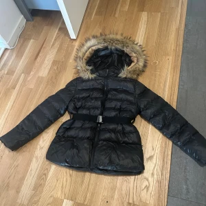 Svart pufferjacka med fuskpäls och bälte - Svart pufferjacka med glansig finish, huva med fuskpäls och markerad midja tack vare ett brett bälte med metallspänne. Jackan har långa ärmar och dragkedja framtill. Perfekt för kalla dagar när du vill hålla stilen.