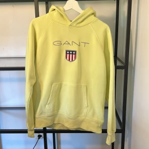 Gul hoodie från GANT med broderad logga - Gul hoodie från GANT med broderad logga och sköld på bröstet. Tröjan har huva, känguruficka och långa ärmar. Perfekt för dig som gillar en sportig och avslappnad stil. Tillverkad i mjuk bomull för extra komfort. Nypris 600 kr.