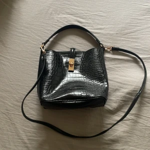Svart axelväska med krokopräglad yta - Snygg svart axelväska med krokopräglad yta och guldfärgat lås. Väskan har både handtag och justerbar axelrem, vilket gör den enkel att bära. Stilren och rymlig insida, perfekt för att få plats med allt du behöver. Materialet är glansigt och ger en lyxig känsla. En liten liten defekt se sista bilden❤️