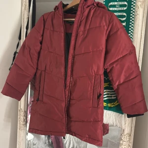 Röd pufferjacka från Take Off - Röd pufferjacka från Take Off med huva och dragkedja framtill. Jackan har två fickor med dragkedja och quiltad design. Perfekt för kalla dagar och har en schysst, enkel look. Materialet är syntet och jackan är långärmad. Passar perfekt på dig om du har storlek 140