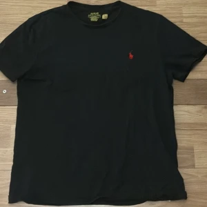 Svart t-shirt Polo Ralph Lauren M - Svart t-shirt från Polo Ralph Lauren i custom slim fit med klassisk röd broderad logga på bröstet. T-shirten har rund hals och korta ärmar, tillverkad i mjuk bomull som känns skön mot huden. Perfekt för dig som gillar stilrena och ikoniska plagg.PRISET ÄR EJ HUGGET I STEN