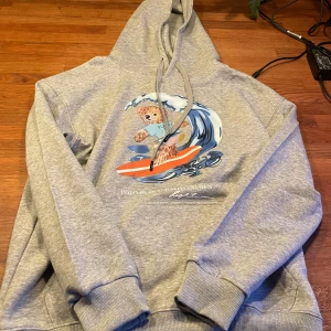 Grå hoodie Polo Bear från Ralph Lauren - Grå hoodie från Ralph Lauren med Polo Bear-tryck där nallen surfar på en våg. Tröjan har huva med snörning, ribbade muddar och klassisk passform. Materialet är mjuk bomullsmix som känns skön mot huden. Perfekt för chill dagar och streetwear-vibe.