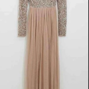 Champagnefärgad långklänning med paljetter - En elegant långklänning i champagnefärg med glittrig paljettöverdel och långa ärmar. Kjolen är i ett mjukt, flytande tyg som ger en drömmig känsla. Perfekt för dig som vill sticka ut med en glamorös look.